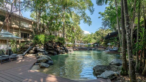 A resort pool area with lounge chairs, a wooden deck, and tropical vegetation.