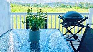 Une table à plateau en verre, agrémentée d’une plante en pot, d’un barbecue, et offrant une vue sur un champ verdoyant et des collines.