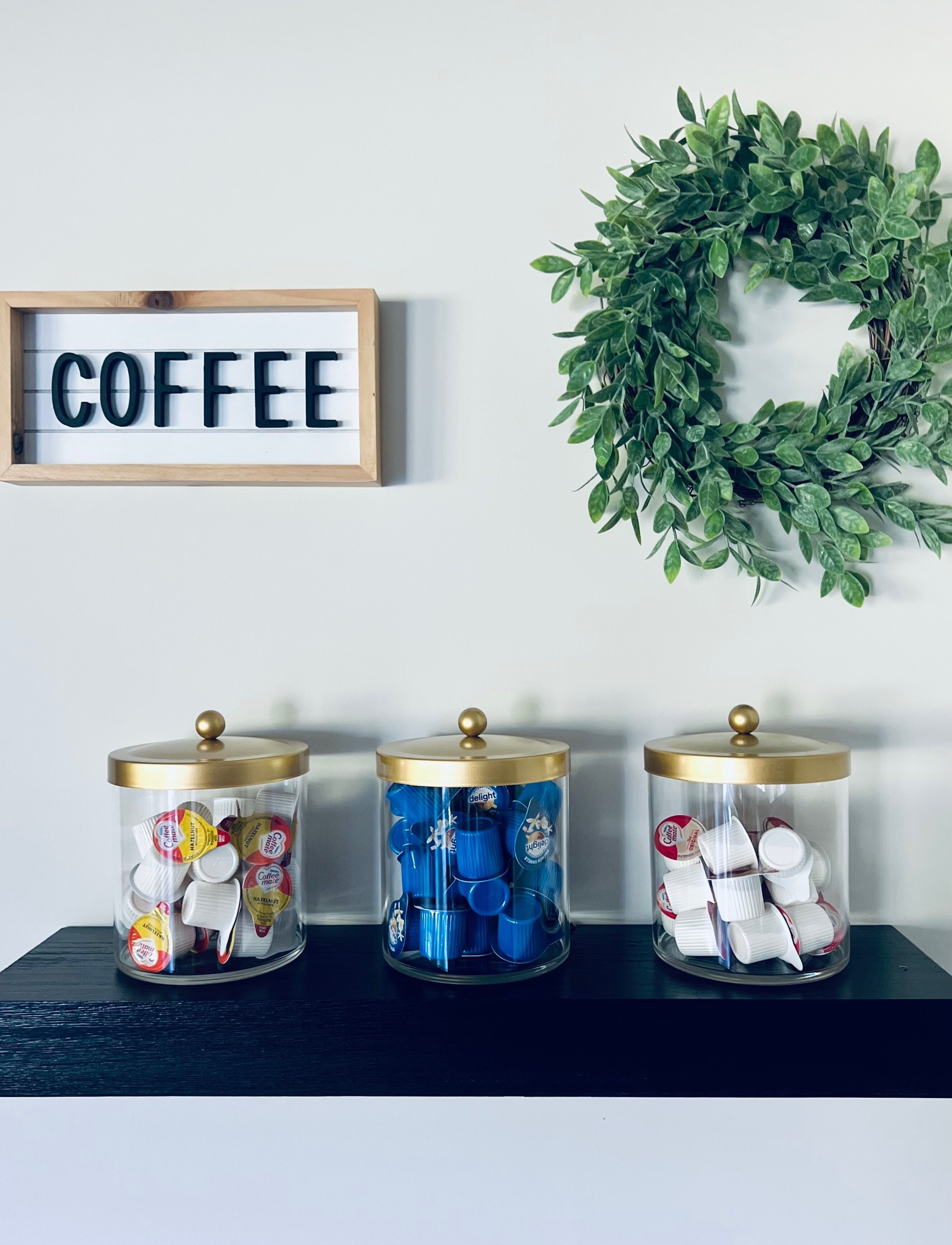 Three glass containers with lids on a shelf, each filled with coffee cups and lids. A wooden sign with the word "COFFEE" is mounted on the wall above the containers.