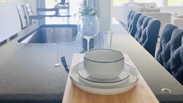 Une cuisine moderne dotée d’un îlot central, d’un coin repas avec des chaises recouvertes de tissu bleu, et offrant une vue sur l’extérieur grâce à de grandes fenêtres.