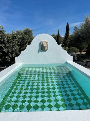 Ein Schwimmbecken mit geometrischem Fliesenmuster, umgeben von weißen Wänden und einer Steinskulptur.