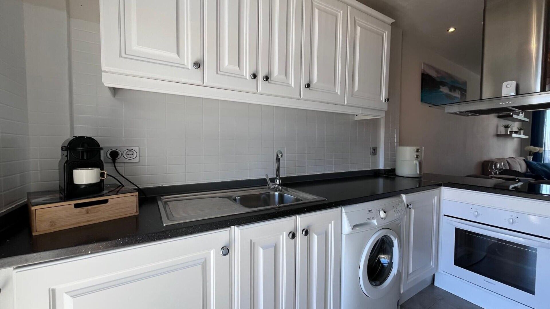 A modern kitchen with white cabinetry, a black countertop, a coffee maker, a washing machine, and an oven.