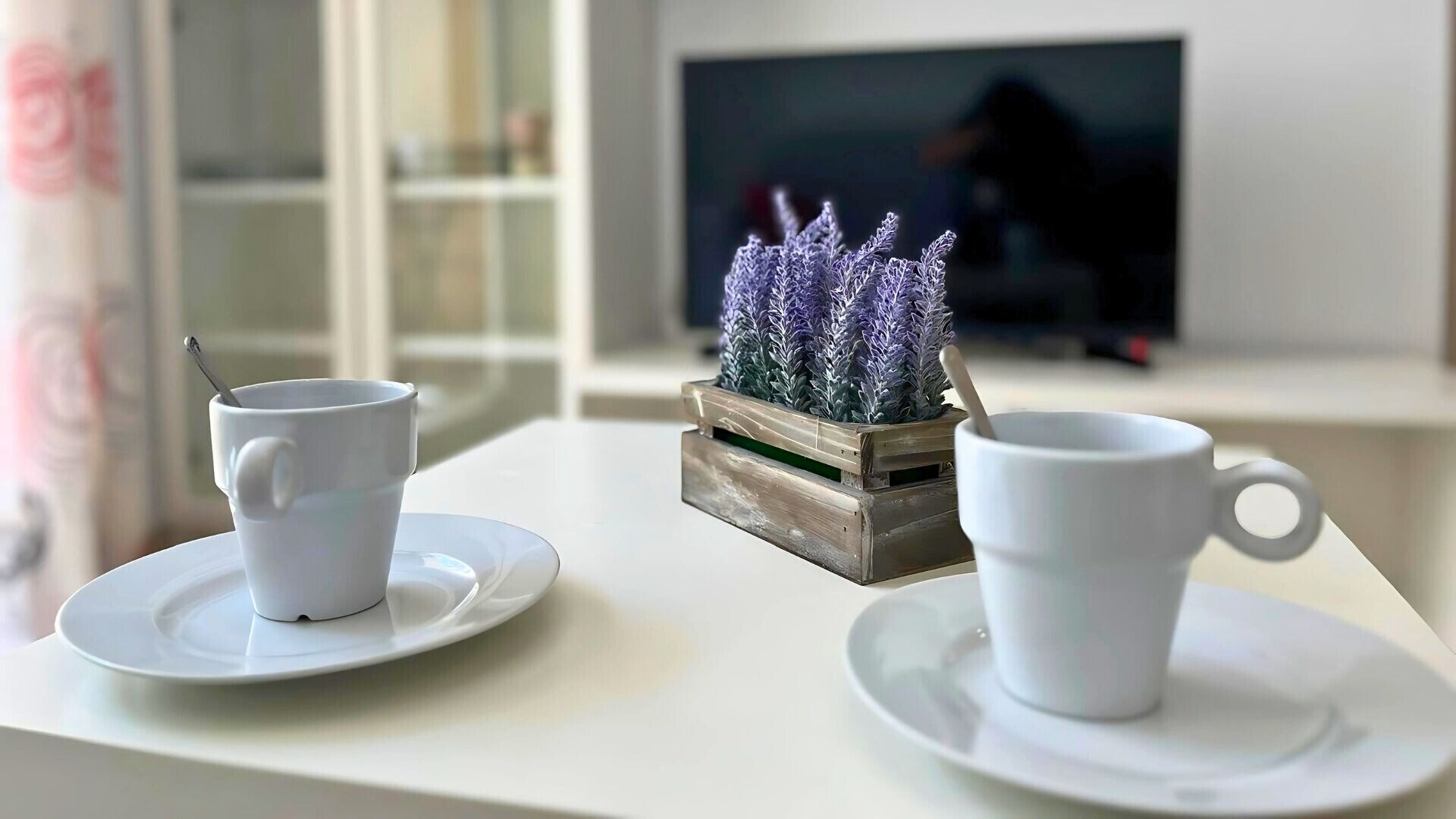 Une table blanche avec deux tasses et soucoupes blanches, un petit pot de fleurs en bois orné de fleurs violettes, et un téléviseur flou en arrière-plan.