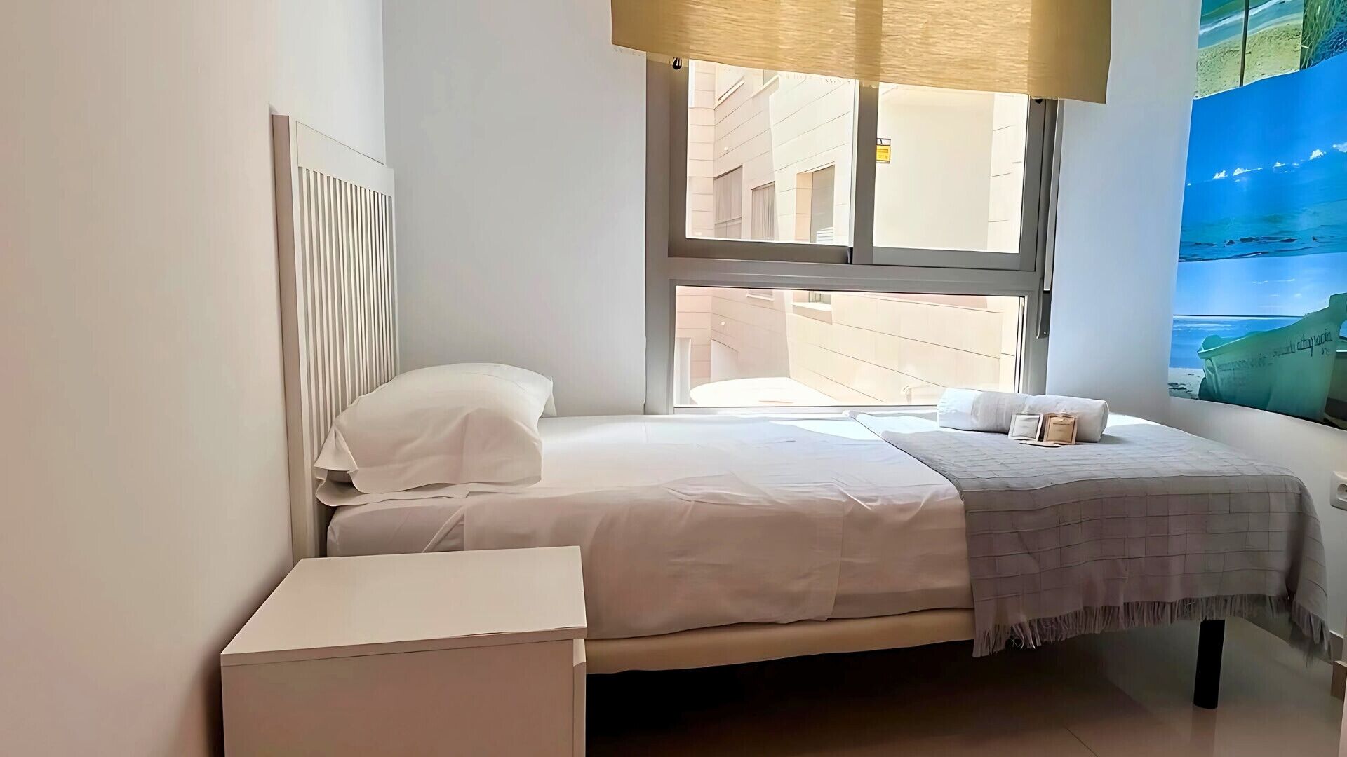 A bedroom with a bed, a bedside table, a window with a view of buildings, and a wall with a mural.