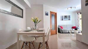 A modern living room with a dining area, featuring a white table set for two, a sofa with red cushions, and a large window with curtains. - Bright flat 50m from the beach with swimming pool and car park (El Campello)