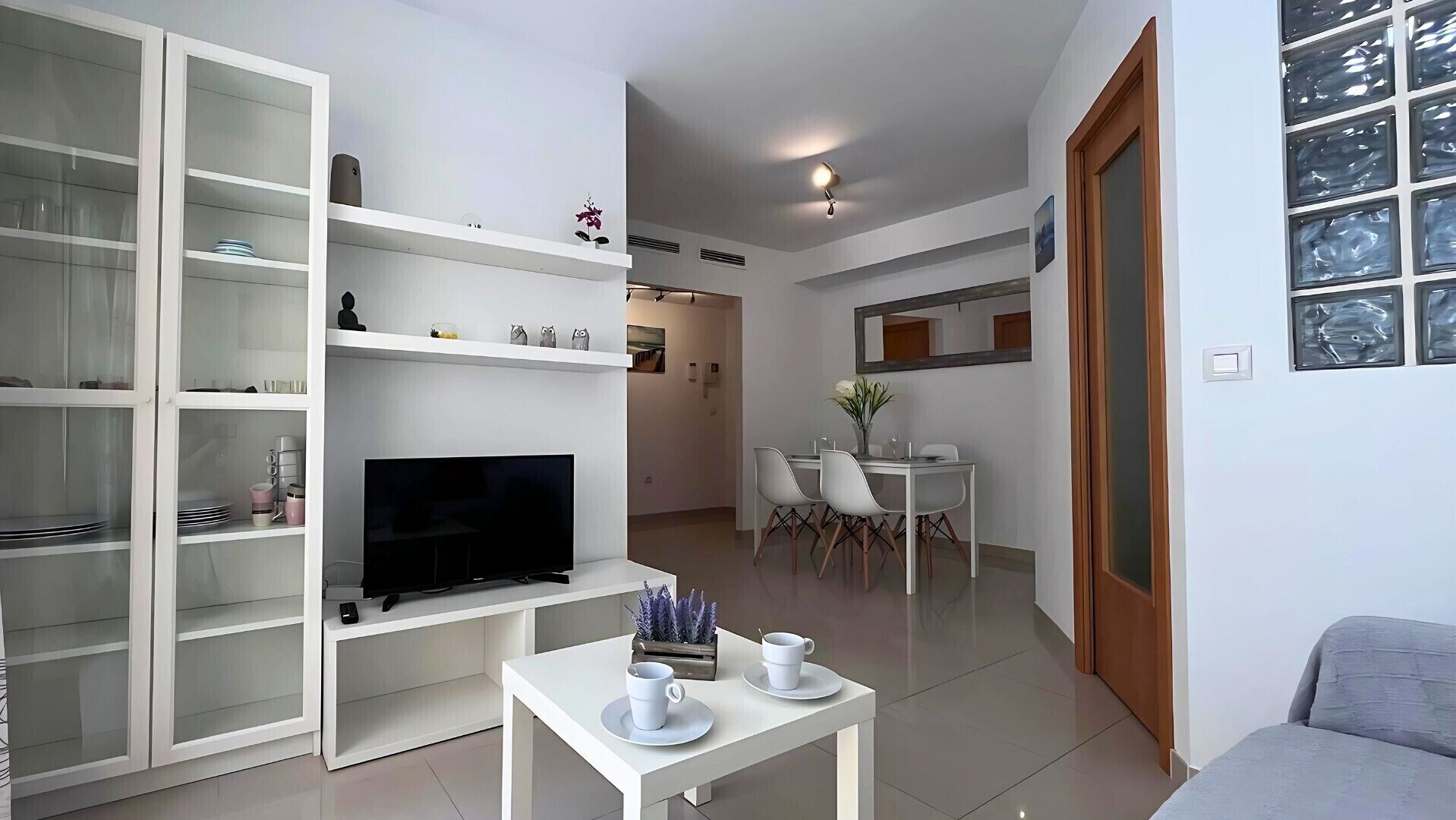 Una sala de estar moderna con un televisor de pantalla plana, muebles blancos y una zona de comedor con sillas y una mesa.