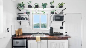 A modern kitchen with white cabinets, a stainless steel refrigerator, a microwave, and a coffee maker. There are potted plants on shelves above the counter and a window with a view of trees. - Exuma Studio w/ King Bed • Ideal for Couples or Solo Travel • Near George Town (George Town)