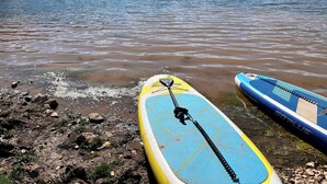 Una tabla de paddleboard sobre una orilla rocosa junto a un lago con colinas al fondo.