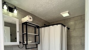 Un baño con lavamanos, inodoro y cortina de ducha blancos. Hay un espejo sobre el lavamanos y un perchero en la pared.