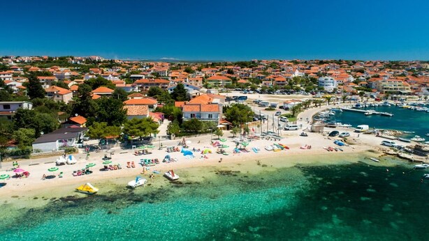 Una vista aerea di una località costiera con spiaggia, acque cristalline turchesi e un porto turistico.