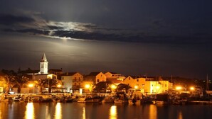 Una cittadina costiera di notte, con una torre imponente, edifici sul lungomare e barche ormeggiate nel porto turistico.