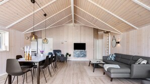 A spacious living area with a high ceiling, wooden walls, a dining table with chairs, a grey sofa, and a flat-screen TV mounted on the wall. - Nice home in Jerup with sauna (Jerup)