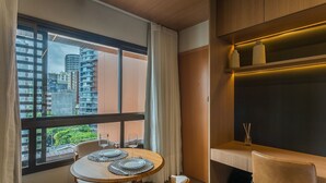 Un coin repas avec une table dressée pour deux personnes, une fenêtre avec des rideaux et une vue sur des immeubles de grande hauteur.
