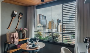 A room with a dining table set for two, a window with blinds, and a view of city buildings. - Units in Building With Swimming Pool in Barra-ssa - EUB (Salvador)