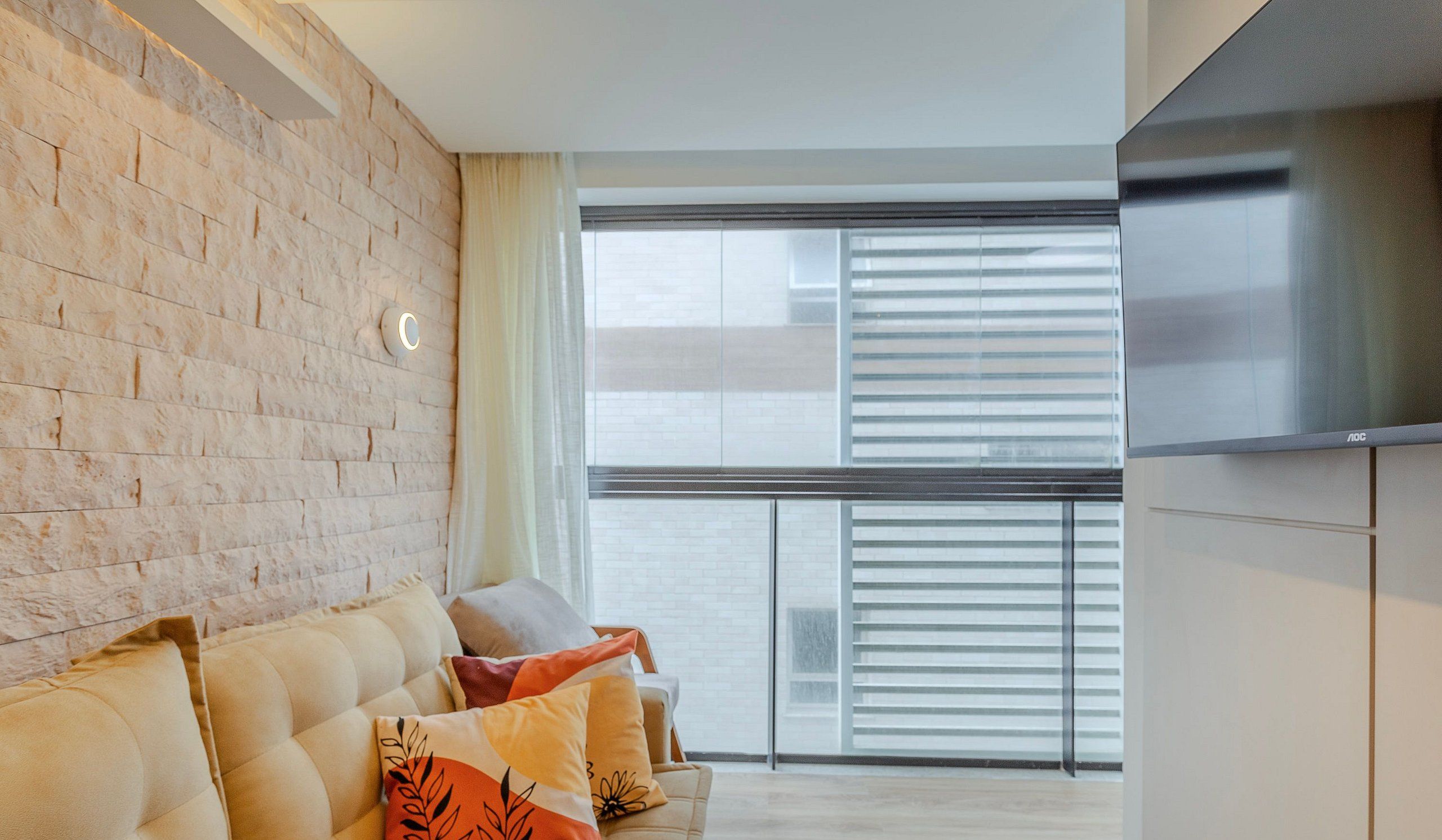 A modern living room with a brick wall, a sofa with cushions, a large window with blinds, and a flat-screen TV mounted on the wall.