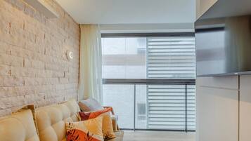 A modern living room with a brick wall, a sofa with cushions, a large window with blinds, and a flat-screen TV mounted on the wall.