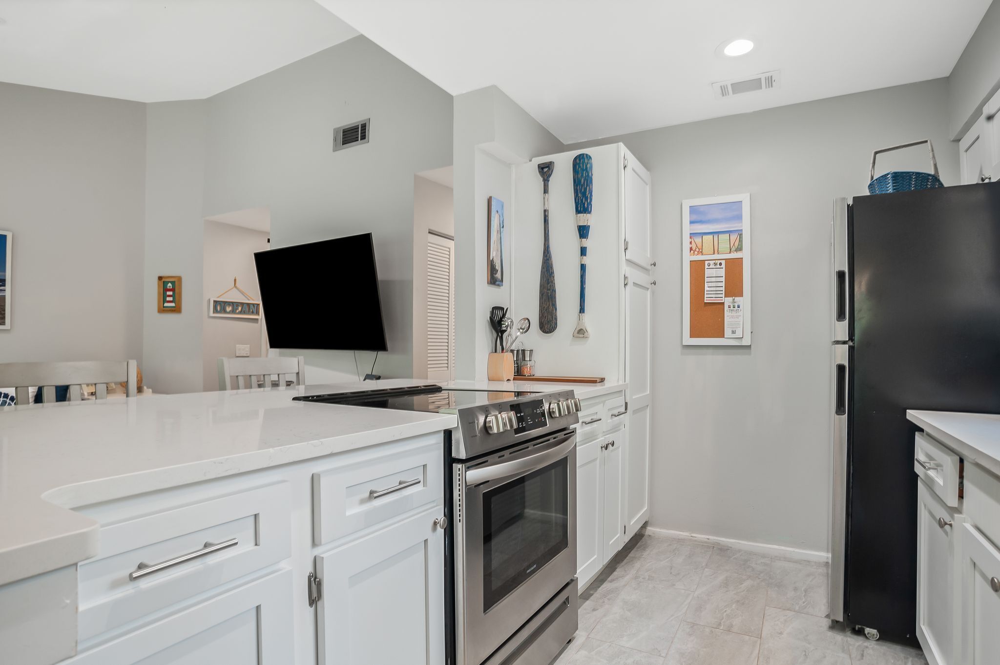 A modern kitchen with white cabinetry, a stainless steel stove, a black refrigerator, and a flat-screen TV mounted on the wall.