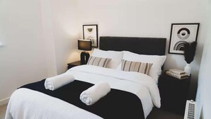 Una cama bien hecha con ropa de cama blanca y dos almohadas, una mesita de noche con lámpara y dos cuadros enmarcados en la pared.