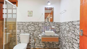 A bathroom with a stone wall, a toilet, a sink with a mirror, and a shower area. - Beautiful Studios in Campos do Jordao - VAE (Campos do Jordão)