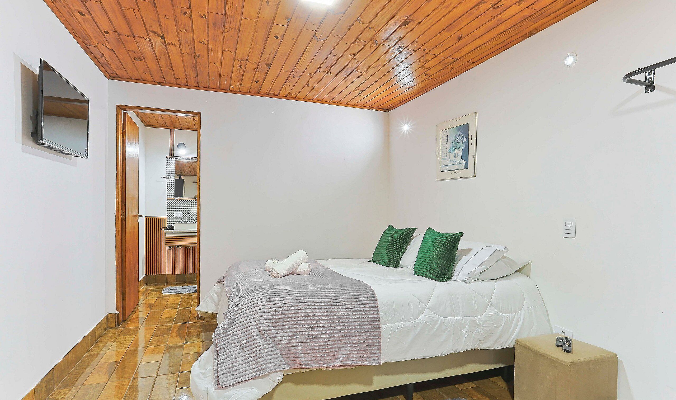 A bedroom with a wooden ceiling, a bed with white bedding and green pillows, a bedside table with a phone, and a small bedside lamp.