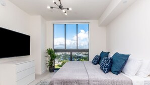 A modern bedroom with a large bed, a flat-screen TV, a potted plant, and a view of a cityscape through large windows.