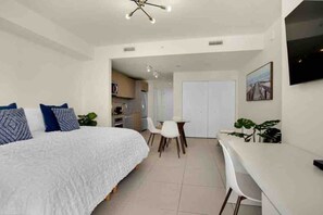 Un dormitorio con cama, escritorio, sillas y una kitchenette al fondo.