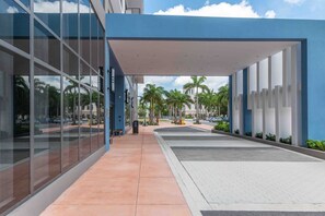 A modern building with large glass windows, a covered walkway, and palm trees in the background. - Modern 1BR Downtown Doral | Walk to Shops & Dining (Doral)