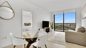 A modern living room with a glass dining table, white chairs, a sofa, a TV, and a balcony with a city view. - Modern 1BR Downtown Doral | Walk to Shops & Dining (Doral)