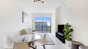 A modern living room with a glass dining table, white chairs, a sofa, a TV, and a large window with a city view.