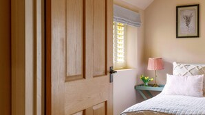 A bedroom with a wooden door, a window with striped valance, a bedside lamp, a framed botanical print, and a bed with white linens. - Lemon Tree Cottage - Luxury Cottages (Painswick)