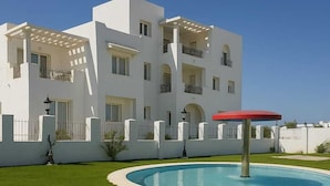 A modern white building with a swimming pool, a red umbrella, and a well-maintained lawn. - Résidence Nesrine (Hammamet)