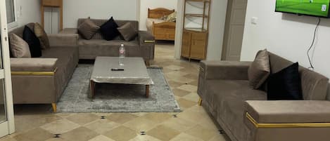 A living room with a sofa set, a coffee table, a TV mounted on the wall, and a patterned floor.