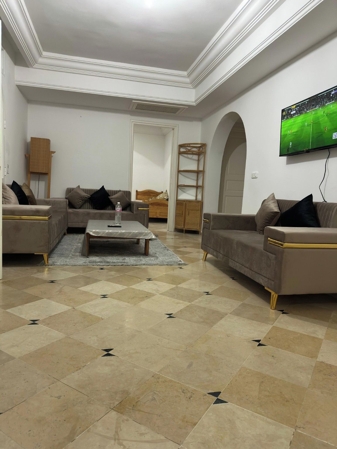 A living room with a checkered floor, sofas, a coffee table, a TV, and a bed in the background.