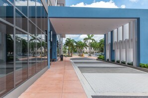 Un bâtiment moderne doté de grandes fenêtres en verre, d’une allée couverte et offrant une vue dégagée sur les palmiers et un parking.