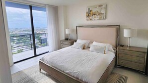 Une chambre moderne avec un grand lit, deux tables de chevet avec des lampes, une vue sur la ville depuis un balcon et un tableau accroché au mur.