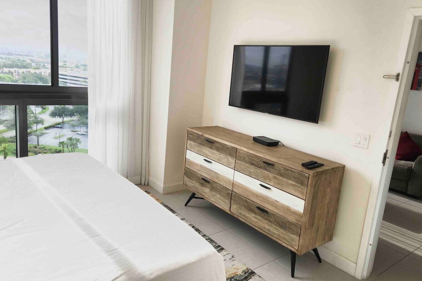 Ein Schlafzimmer mit einem Bett, einer Holzkommode, einem an der Wand montierten Fernseher und Blick auf eine Stadtlandschaft durch ein Fenster.