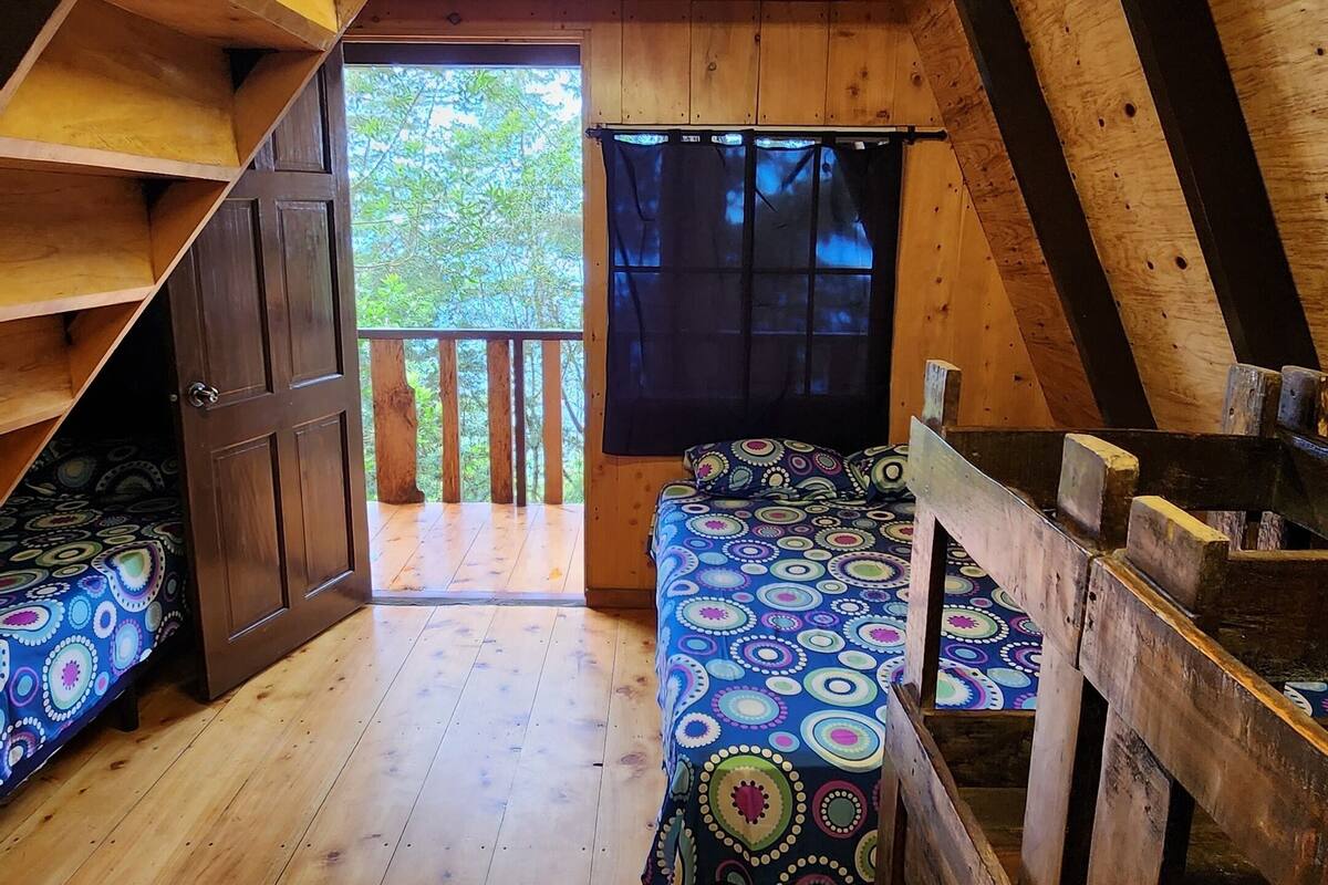 A cozy attic bedroom with a sloped ceiling, wooden beams, a bed with a colorful patterned bedspread, and a window with a view of greenery.