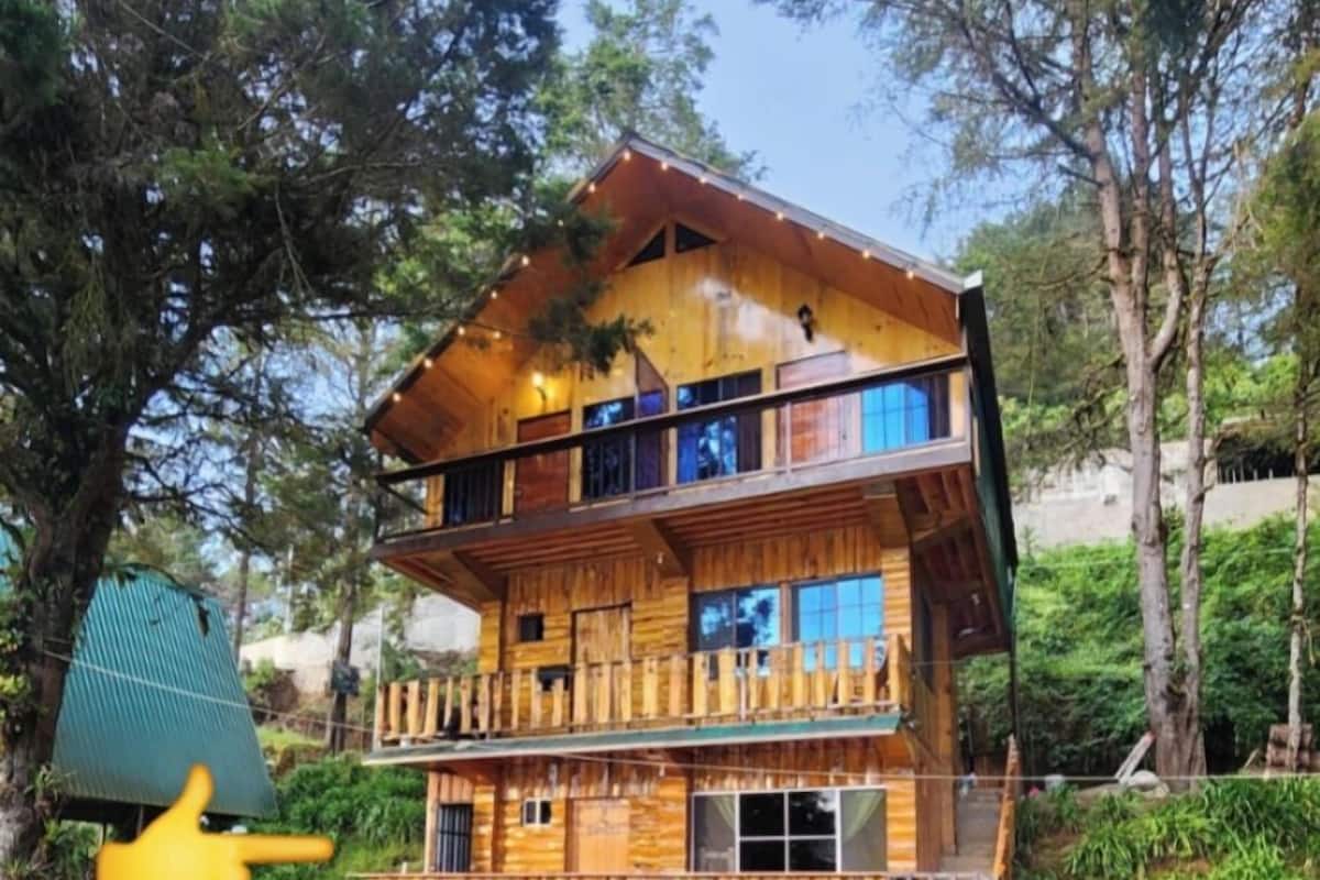 A multi-story wooden cabin with a balcony on the upper floor, surrounded by greenery.