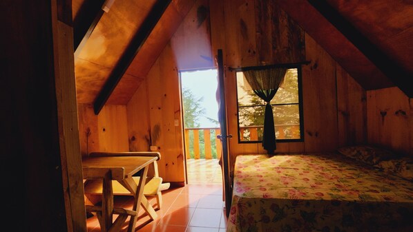 A wooden cabin room with a bed, a table, and a window with curtains.