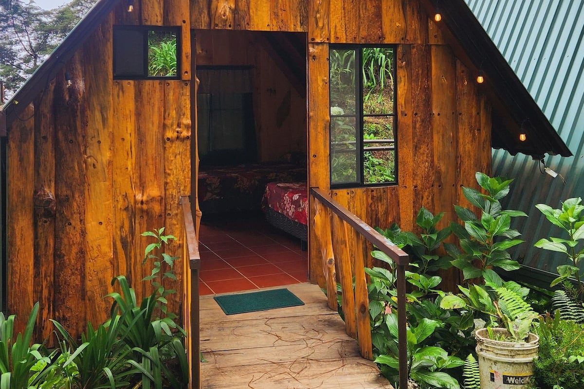 A rustic wooden cabin with a steeply pitched roof, a small porch with a railing, and a door leading inside.