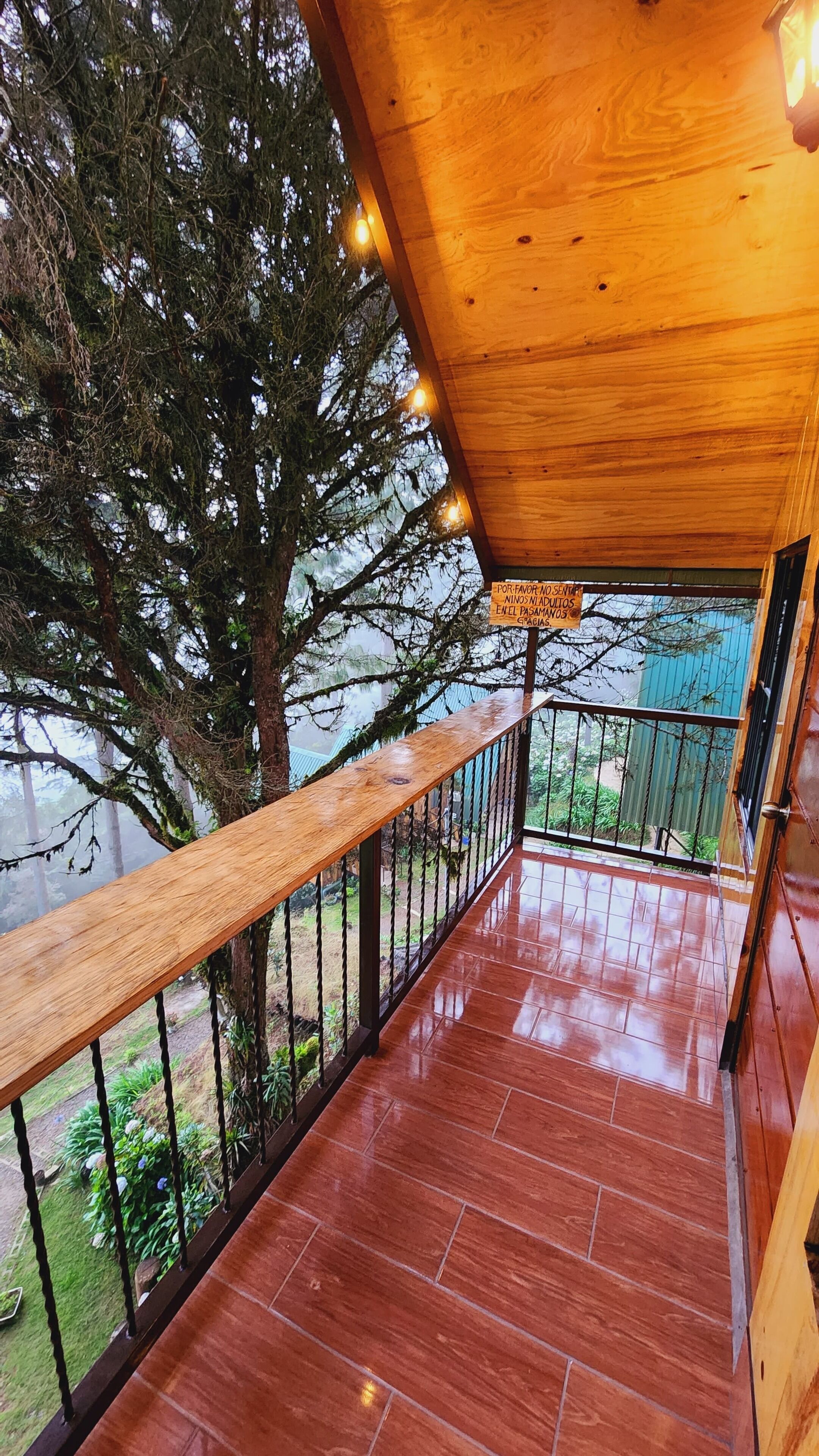 A wooden balcony with a metal railing, a tiled floor, and a view of a tree and greenery.