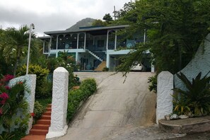 A two-story building with a staircase leading to the entrance, surrounded by lush greenery and a mountain in the background.