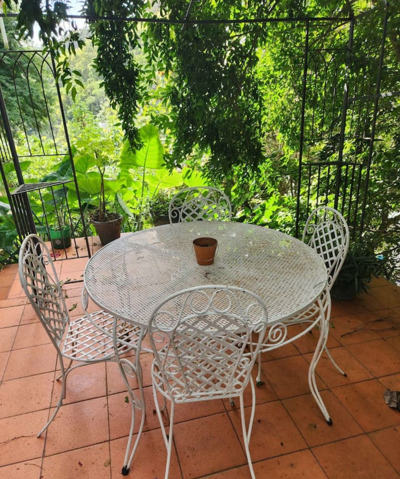 Un tavolo rotondo in metallo con quattro sedie, circondato da piante in vaso, su un patio piastrellato con ringhiera in metallo.