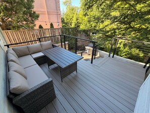 Una terrazza con un divano e un tavolo, circondata da un parapetto e vegetazione.