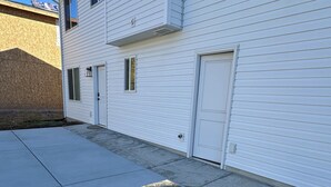 Un foyer extérieur carré, doté d’un dessus en pierre et entouré d’une structure en métal noir, est installé sur une terrasse en béton devant un bâtiment blanc avec une porte de garage.