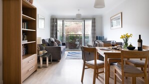 A modern living room with a sofa, dining table, and chairs. There is a bookshelf, a lamp, and a window with a view of the outdoors.