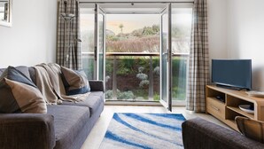 A modern living room with a grey sofa, a blue and white striped rug, a television on a wooden stand, and a sliding glass door leading to a balcony with greenery.