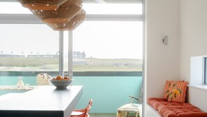 A modern interior with a large window, a dining area with orange chairs, a small table with a bowl of fruit, and a cozy seating area with a cushion.