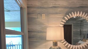 Une pièce avec une grande fenêtre, un miroir fixé au mur, une lampe et une commode en bois.