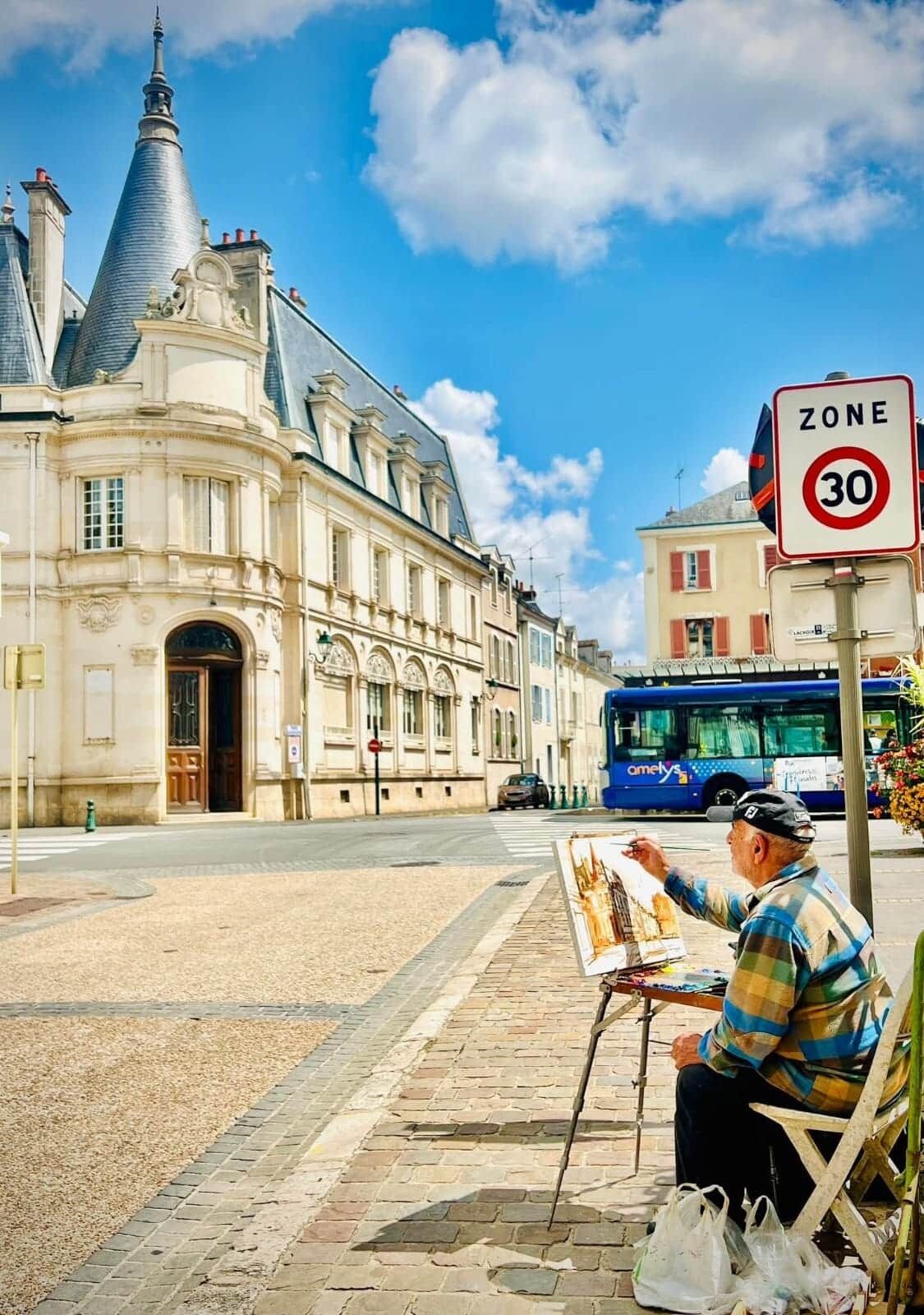 Un artiste de rue est en train de peindre sur un banc devant un bâtiment historique, à côté d’un panneau indiquant une limite de vitesse de 30.